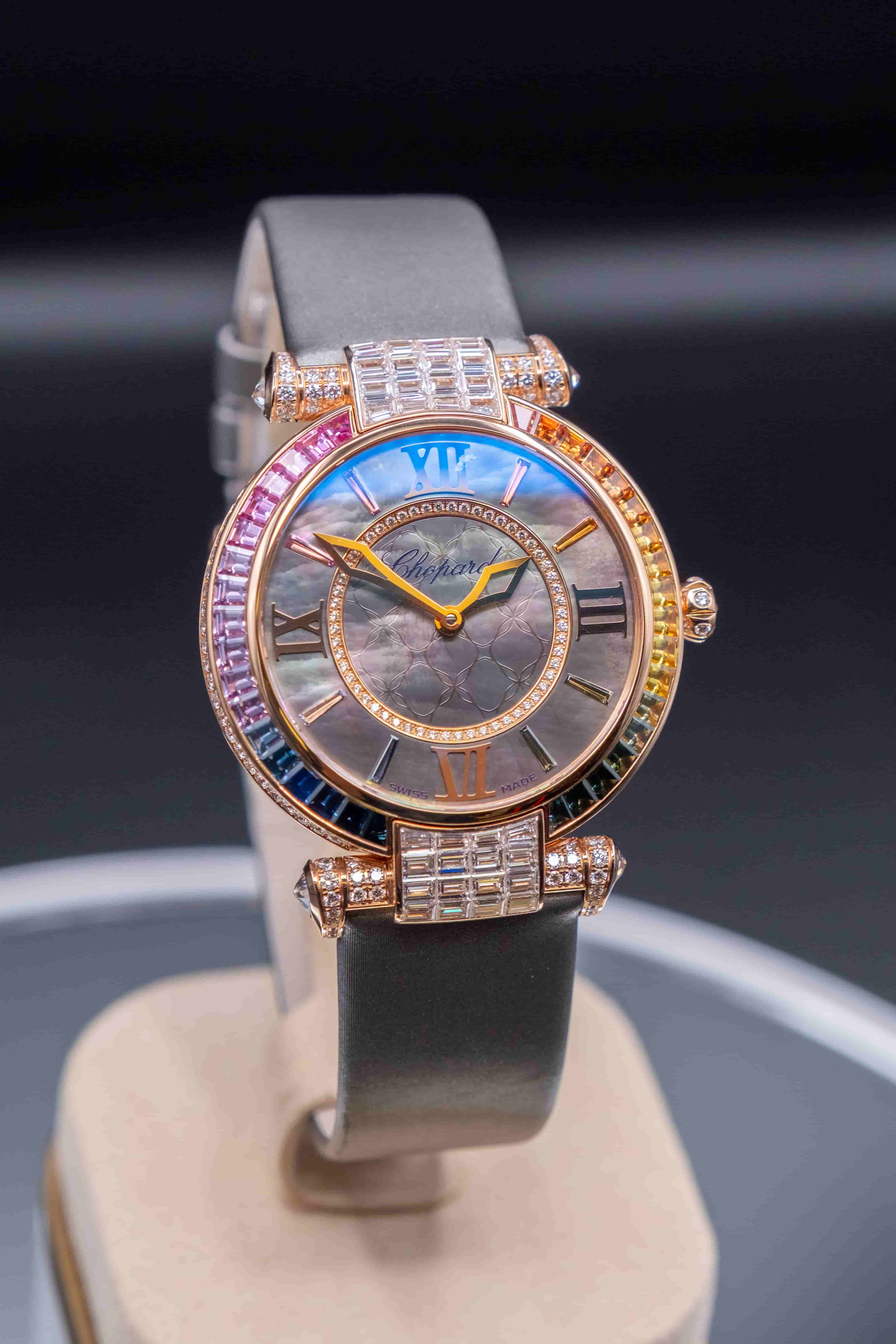 Luxury Chopard Imperiale watch with a rainbow gemstone bezel, dark mother-of-pearl dial, and a grey satin-finish leather strap.